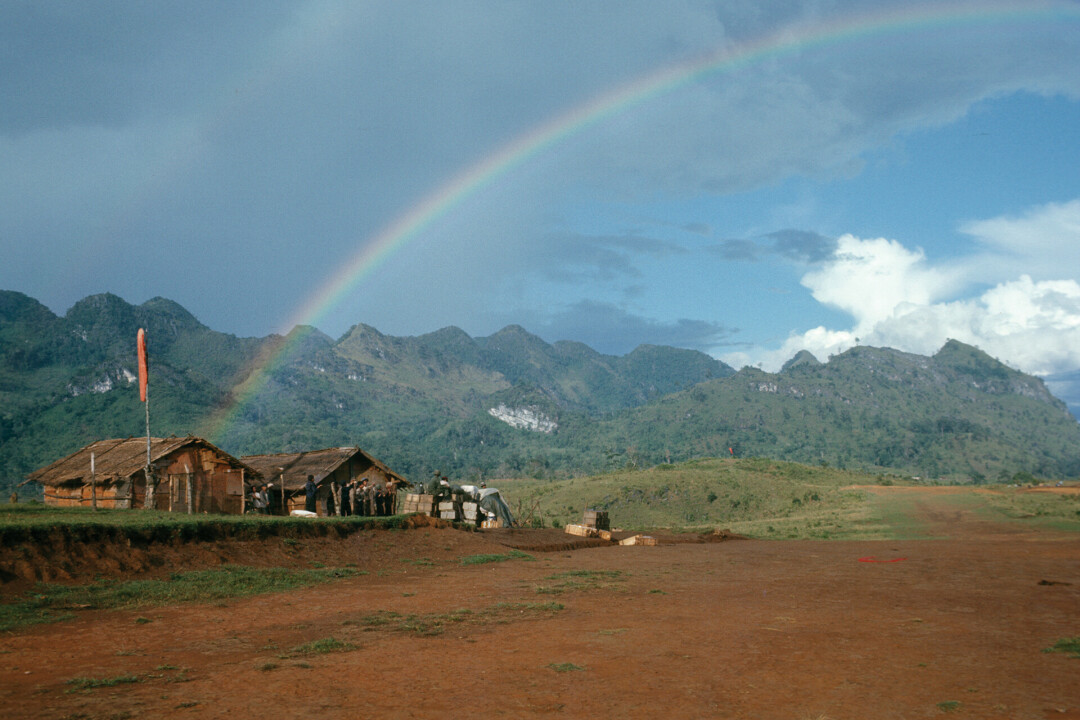 A rainbow cascades across the sky above an airstrip at the Tham Bleung Hmong refugee site. Photographed in October 1969 by Frederic Benson.
